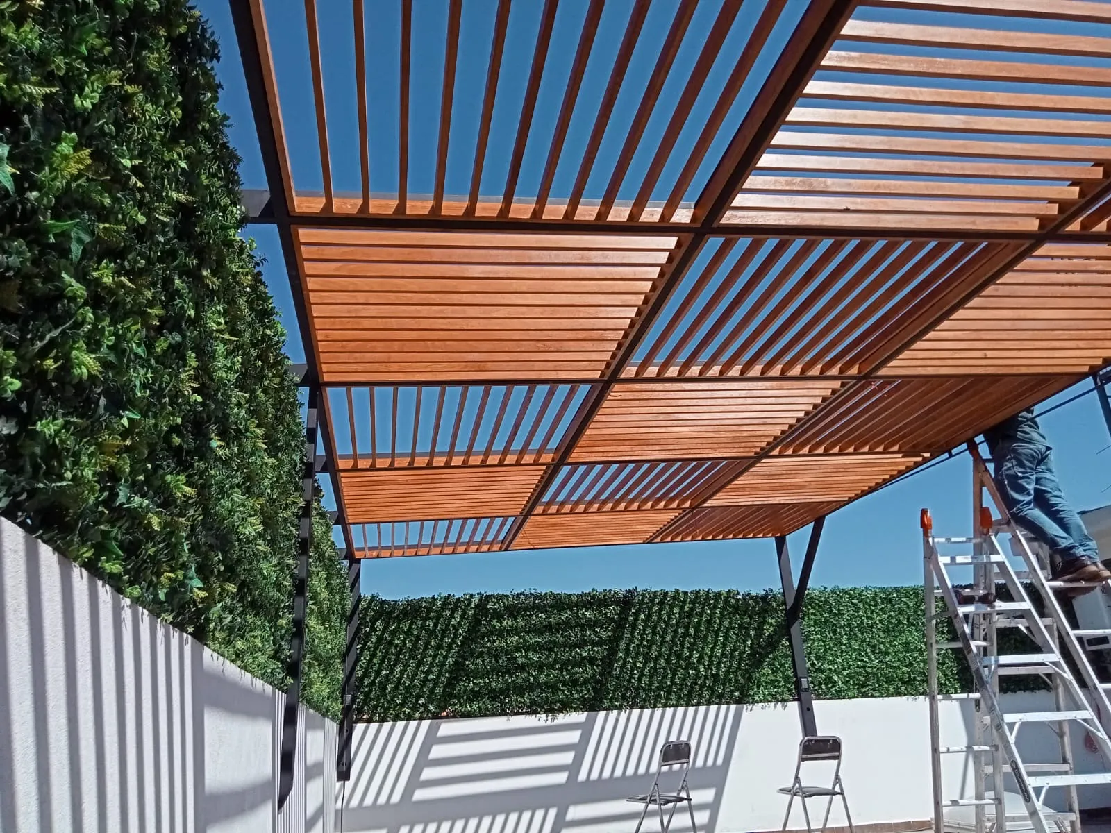 Vista inferior de celosía de madera sobre estructura de acero negro junto a muro verde